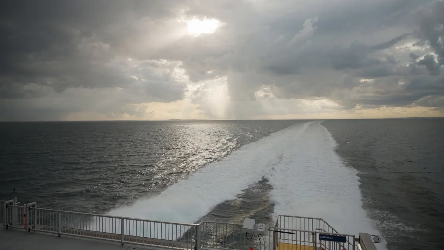 The wake of a ferry running to the horizon under a broken, overcast sky