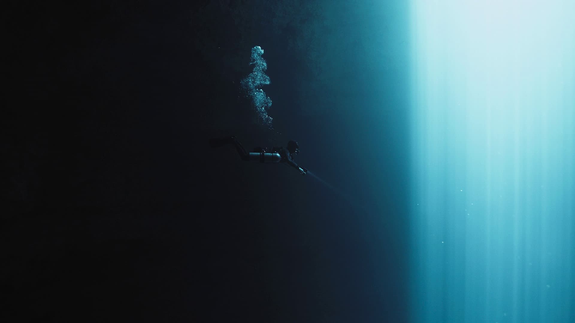A diver in full kit descending into deep blue water, light falling from the surface behind